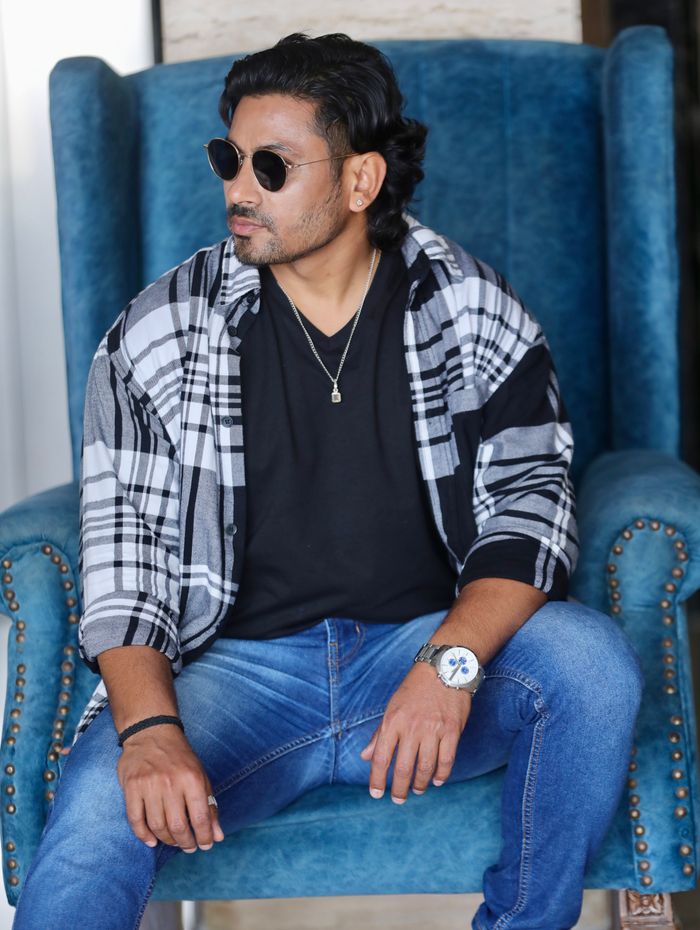 A trendy man in sunglasses sits stylishly in a blue armchair indoors.